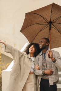 A couple in warm clothing stands together under an umbrella. The woman points to something ahead, and the man is smiling.