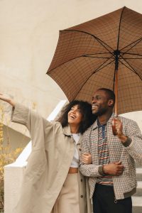 A couple in warm clothing stands together under an umbrella. The woman points to something ahead, and the man is smiling.