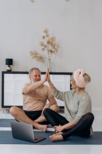 An older couple, wearing workout clothes, sit on yoga mats and high five after a workout in their living room.