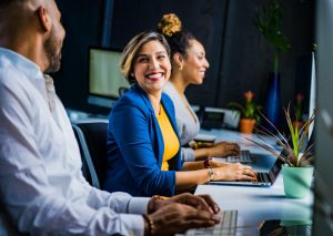 Three coworkers sit at desks and smile up from their computers.