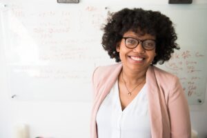 A black business woman, wearing glasses, smiles in front of a whiteboard.